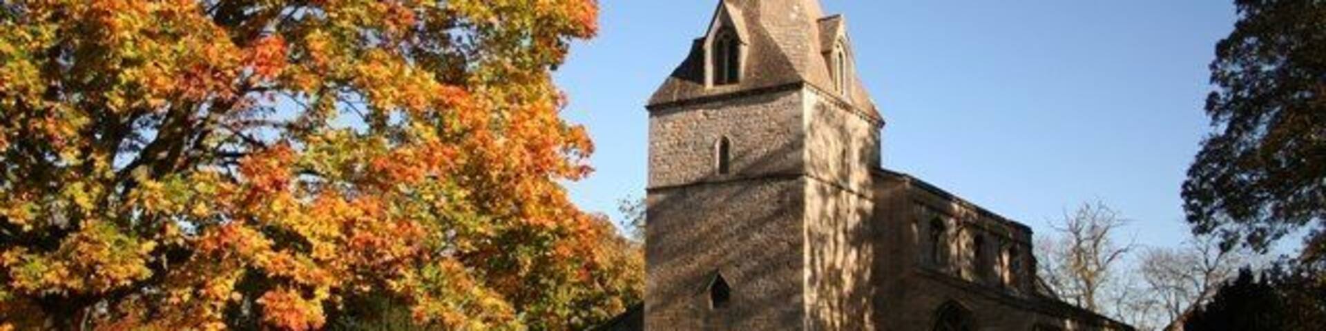 A church with a tall steeple and a cross on top, surrounded by autumn trees and a fenced area.