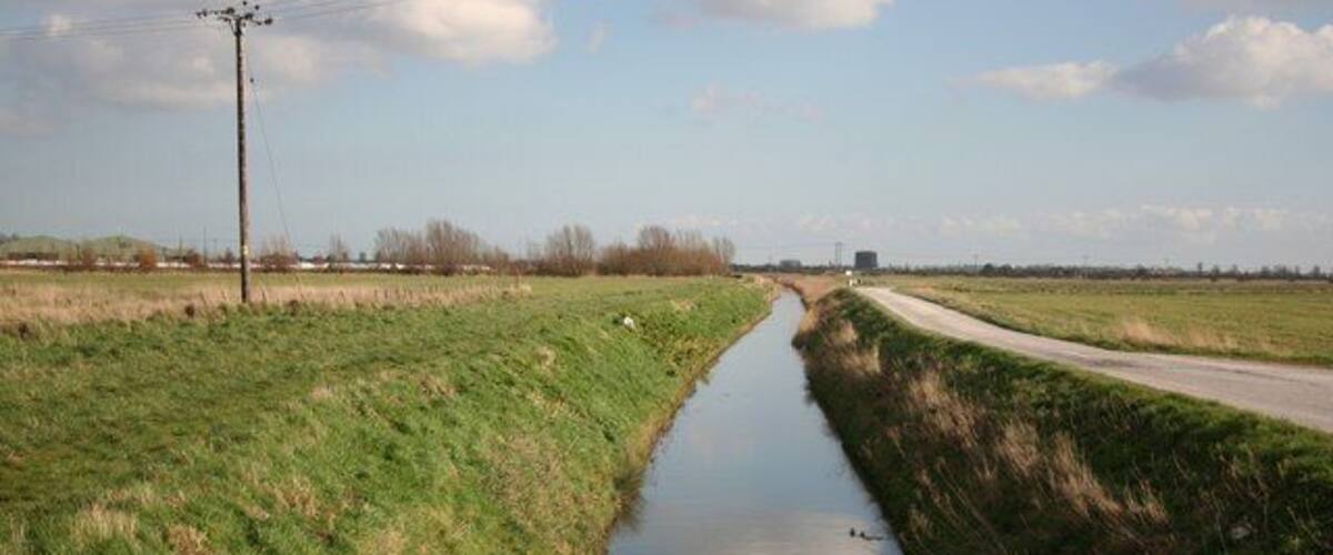 A narrow waterway flanked by grassy fields, a road, and a distant bridge.