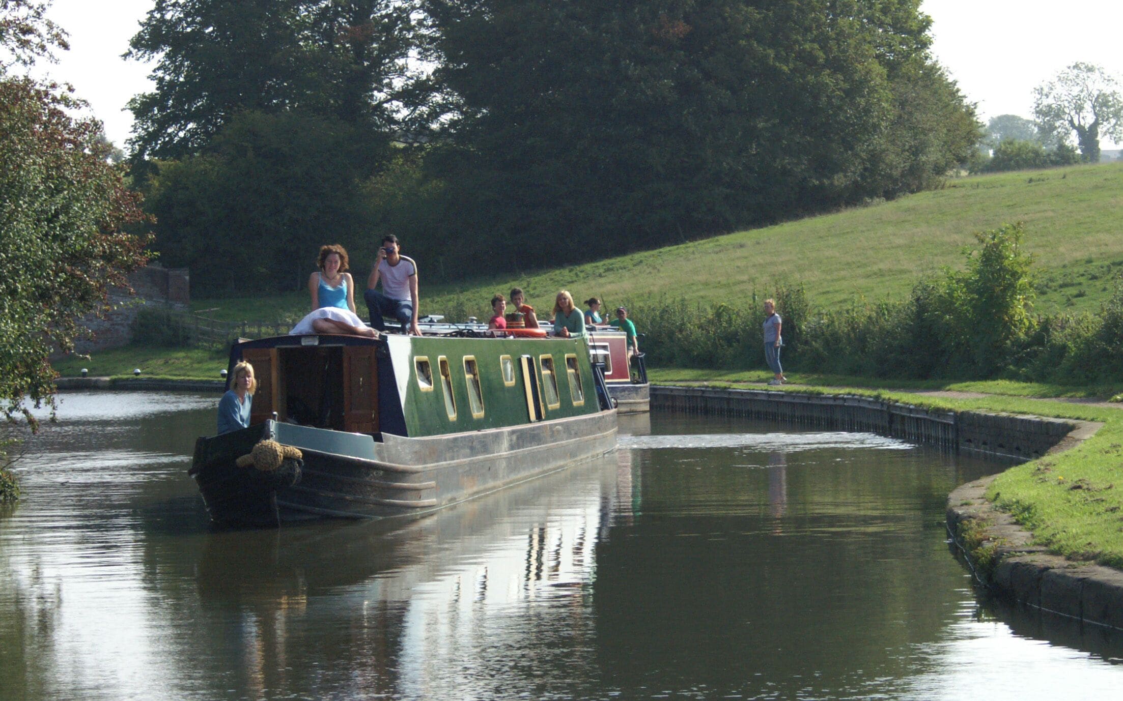 A narrowboat with passengers on a canal surrounded by greenery.