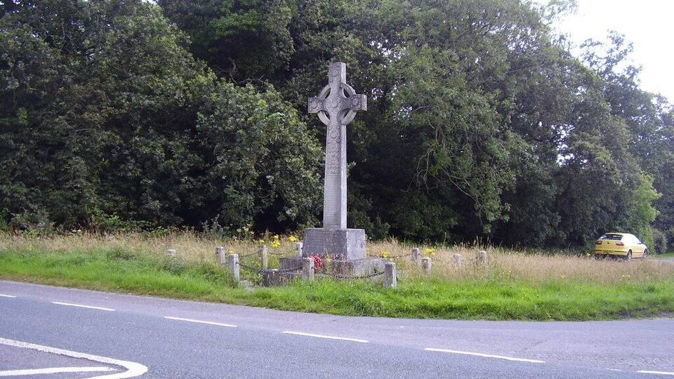 A stone cross monument surrounded by greenery and a road with a yellow car in the background.