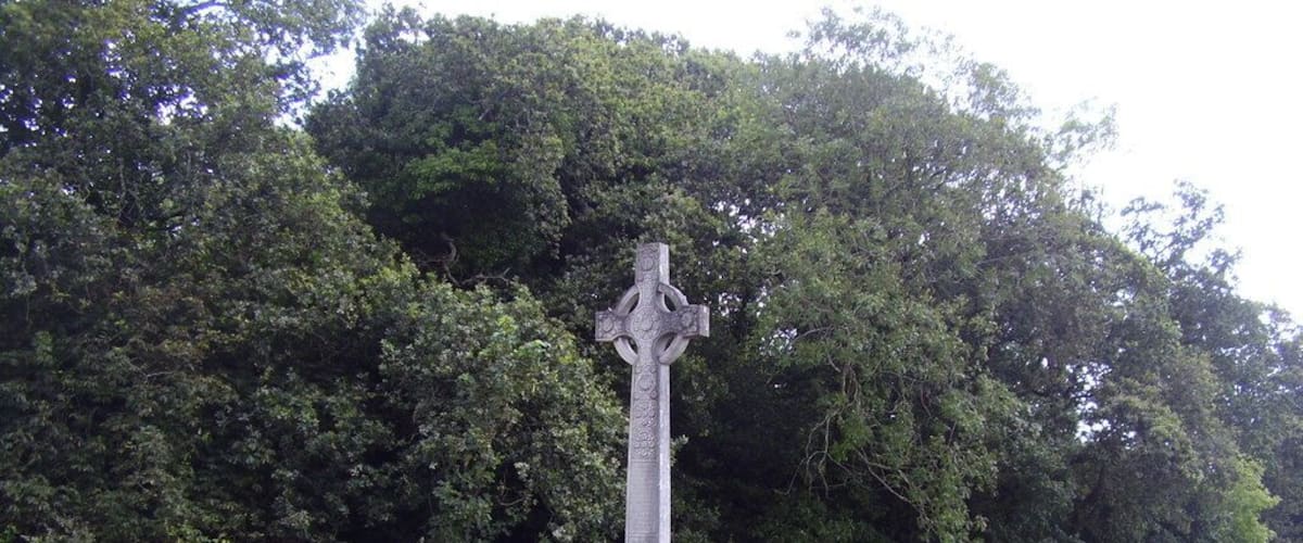 A stone cross monument surrounded by greenery and a road with a yellow car in the background.