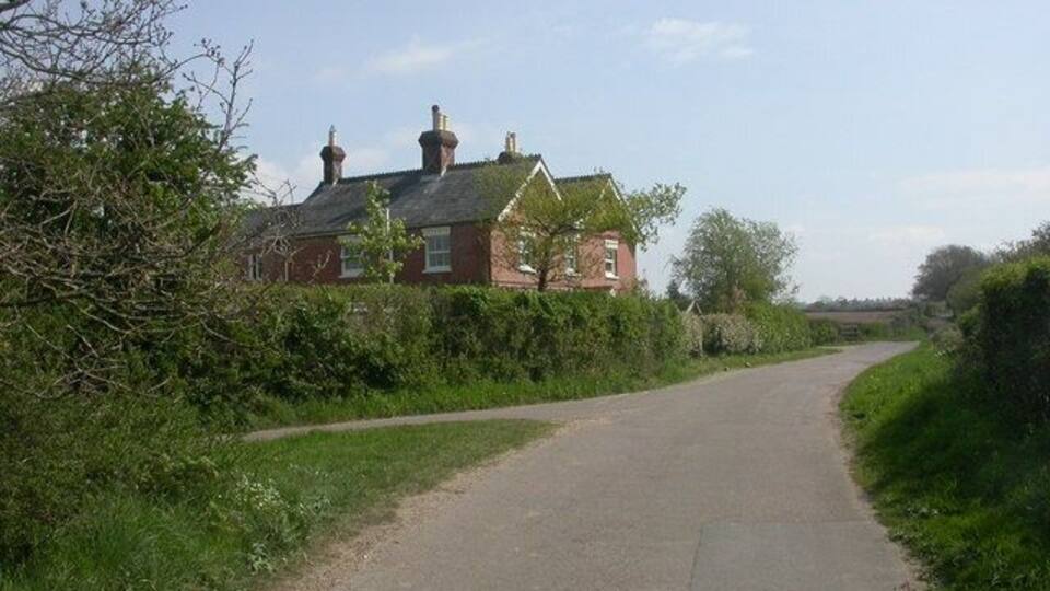 A narrow rural road curving through a green landscape with a red brick house on the left.