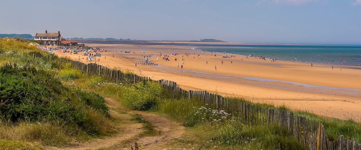 A sandy beach with a wooden fence, a path, and a building in the background.