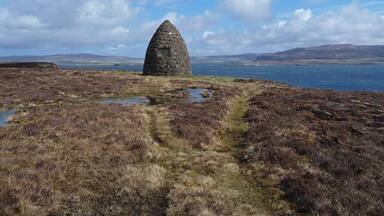 A solitary stone structure on a grassy hill with a lake and mountains in the background.