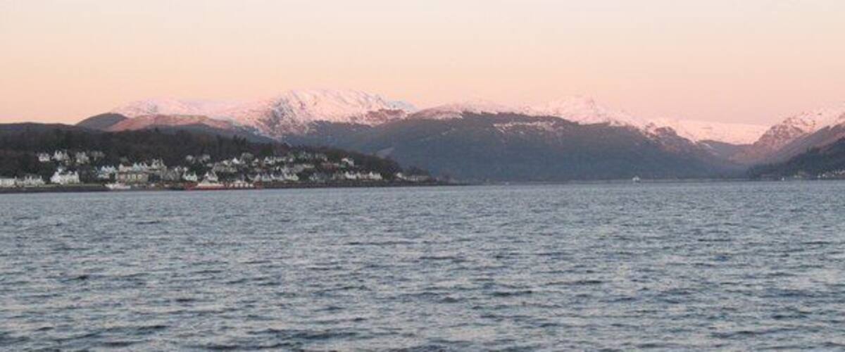 A calm lake with snow-capped mountains in the background and a small settlement on the left.