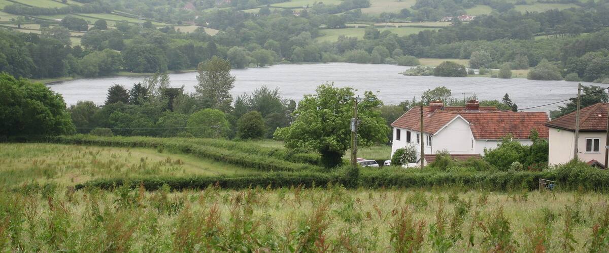 A river flowing through a rural landscape with rolling hills, a white house with a red roof, and lush greenery.