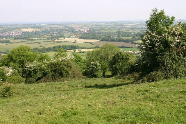 A grassy hill with trees and a view of a valley with fields and a small town.