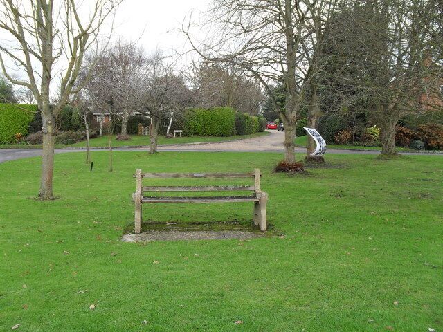 A park with a wooden bench, leafless trees, a sculpture, and a road in the background.