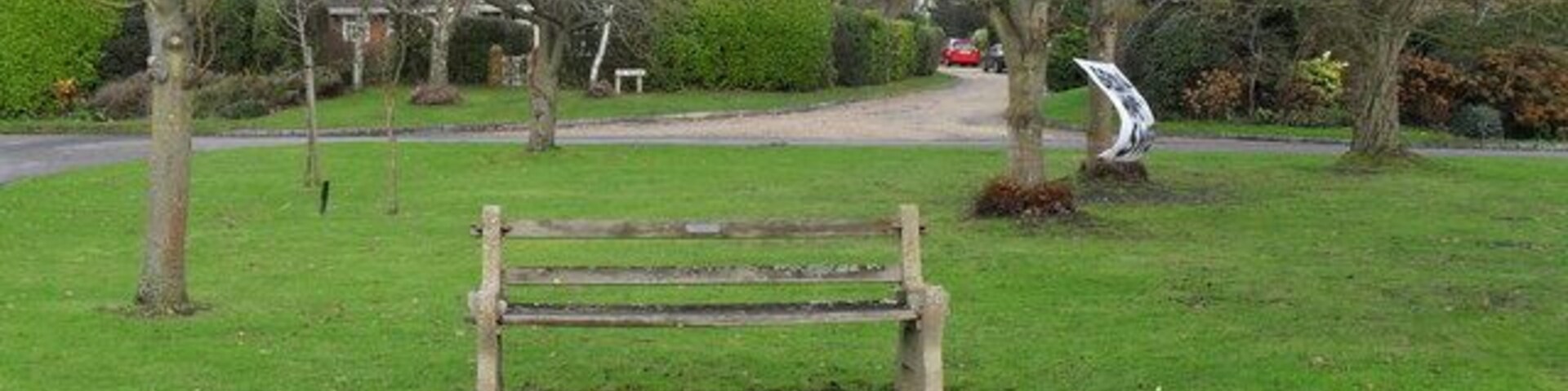 A park with a wooden bench, leafless trees, a sculpture, and a road in the background.