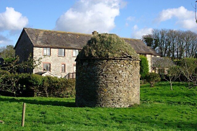 A stone tower in a grassy field with a stone house in the background.