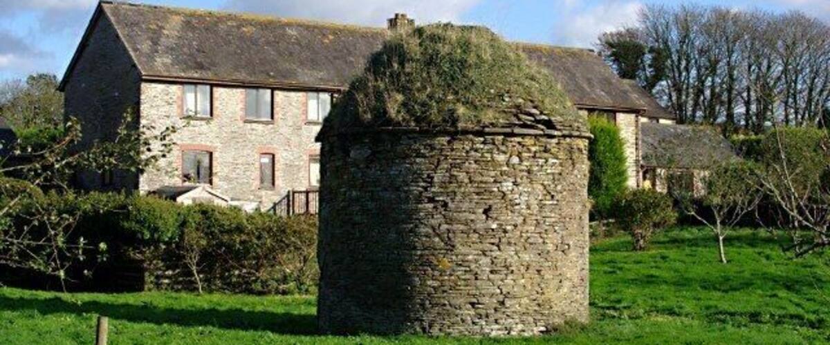 A stone tower in a grassy field with a stone house in the background.