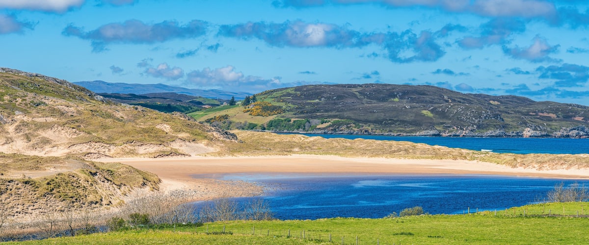 A coastal landscape with a sandy beach, grassy hills, and a clear blue sea.