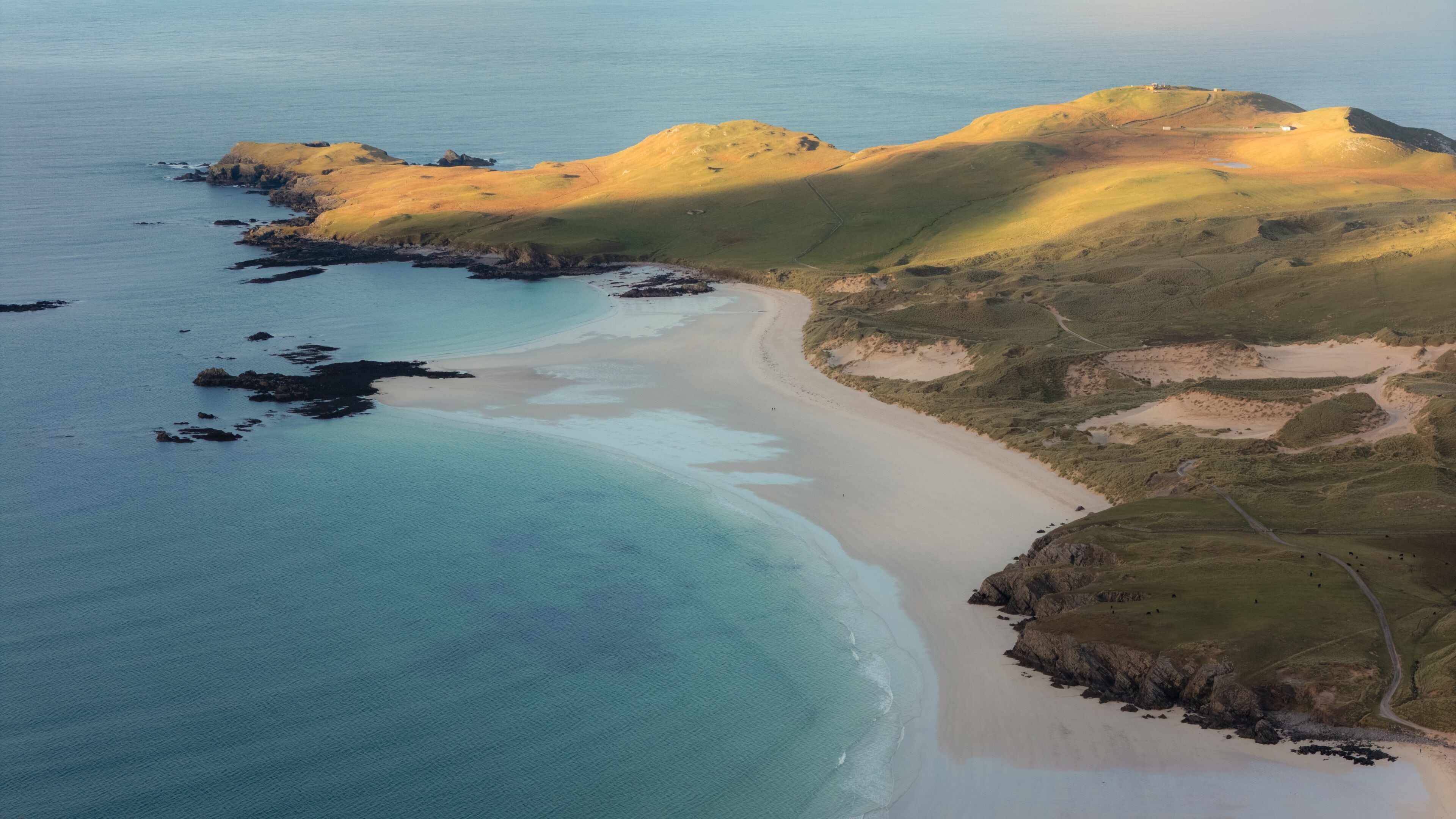 Eine Küstenlandschaft mit Sandstrand, klarem türkisblauem Wasser und sanften Hügeln mit spärlicher Vegetation.