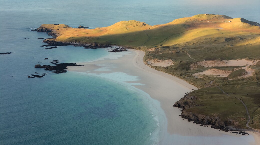 Eine KĂŒstenlandschaft mit Sandstrand, klarem tĂŒrkisblauem Wasser und sanften HĂŒgeln mit spĂ€rlicher Vegetation.