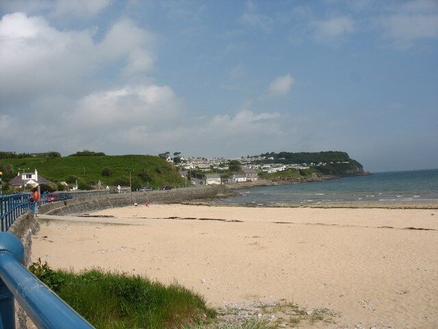A sandy beach with a blue railing, a rocky shoreline, and houses on a hill in the background.