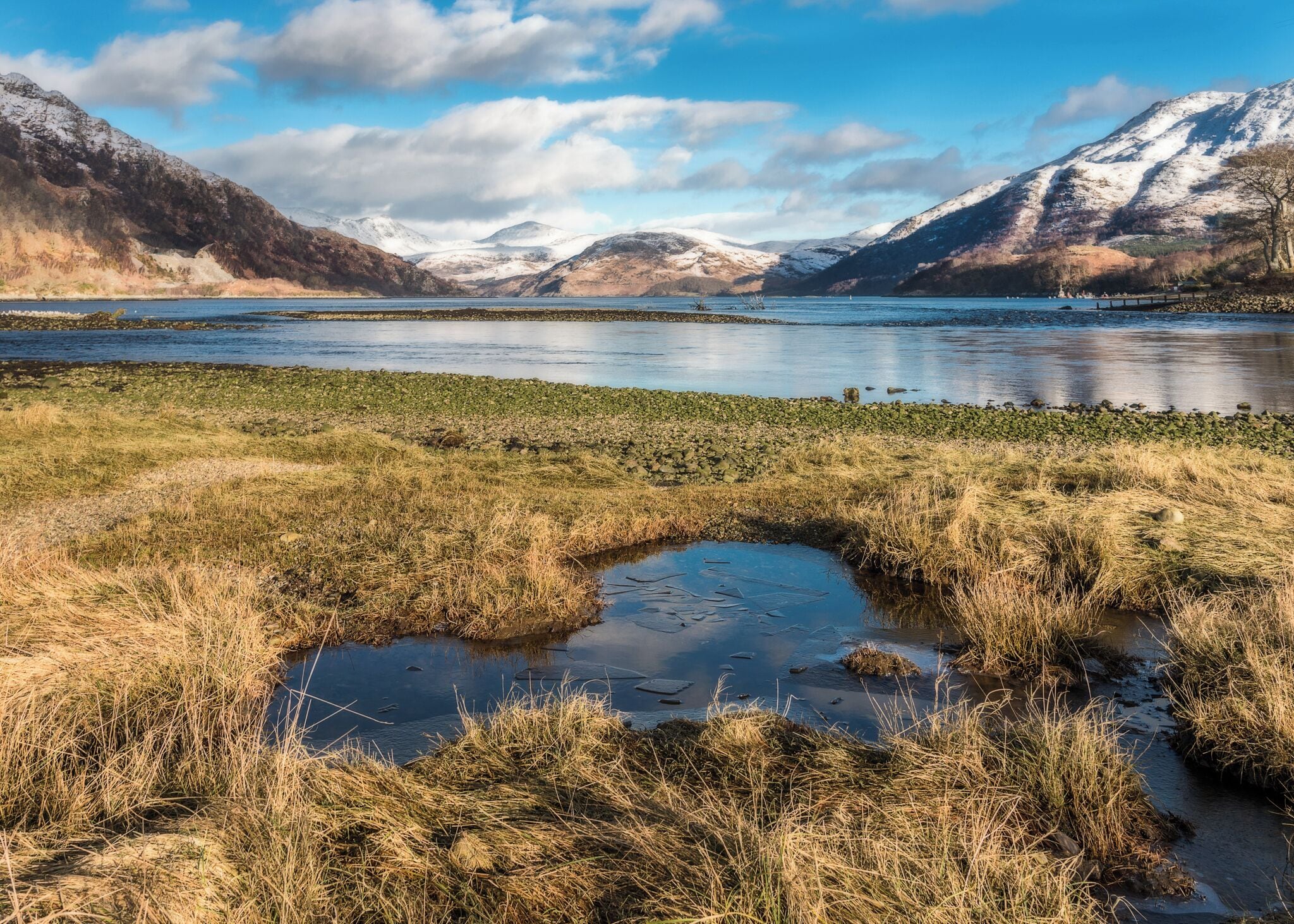 A lake surrounded by mountains with patches of snow, a small pond, and a grassy shoreline.