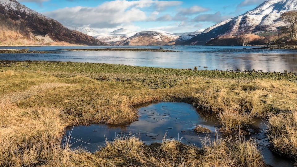 A lake surrounded by mountains with patches of snow, a small pond, and a grassy shoreline.