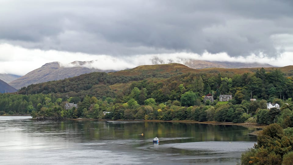 A lake with a boat, surrounded by forested hills and houses.