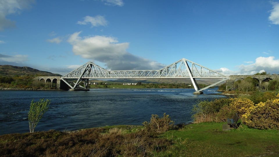 A steel bridge over a river with green hills in the background.