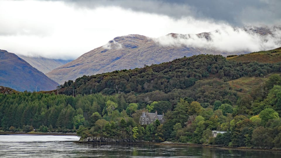 A lake surrounded by mountains, a house on a hill, and dense forest.