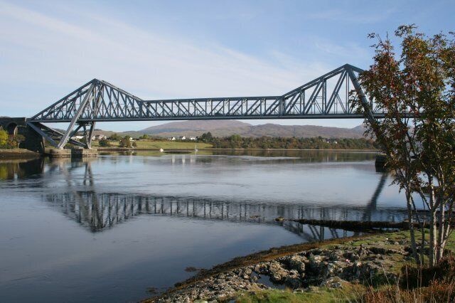 A steel bridge over a calm river with a mountain in the background.