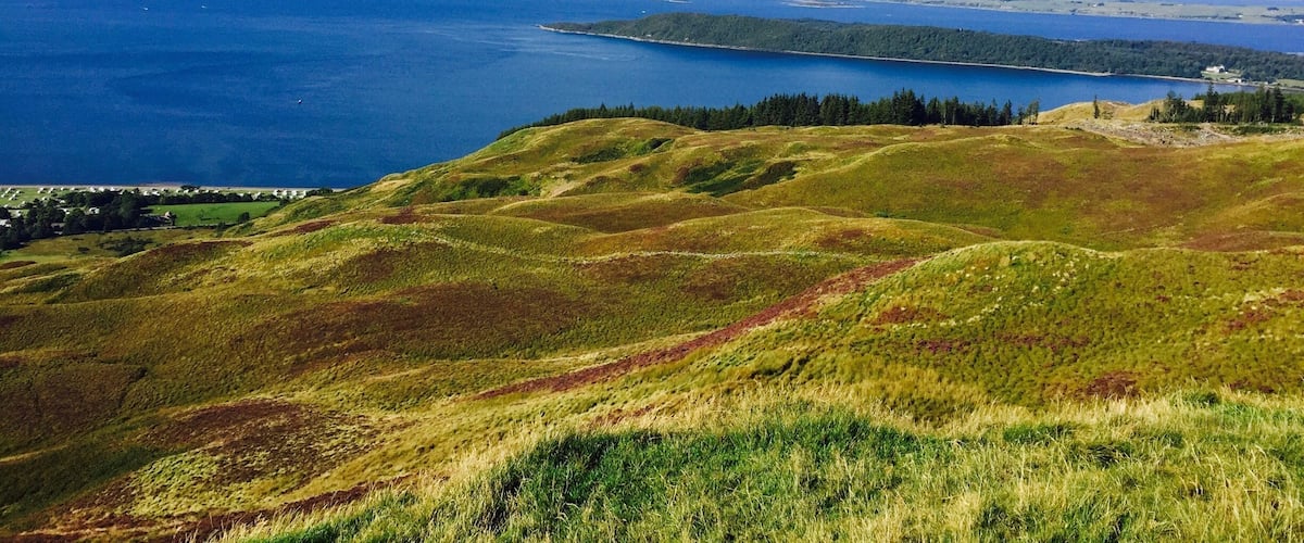 A coastal landscape with rolling hills, a large body of water, and distant mountains.