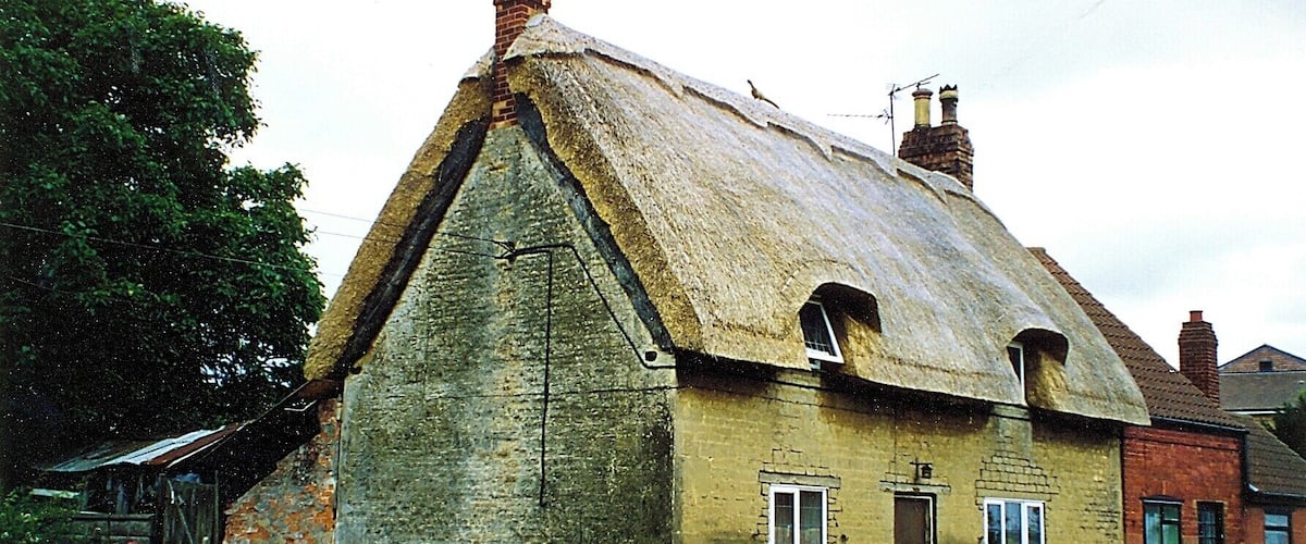 A traditional thatched-roof house with a stone foundation, set in a rural area with a road in the foreground and greenery around.