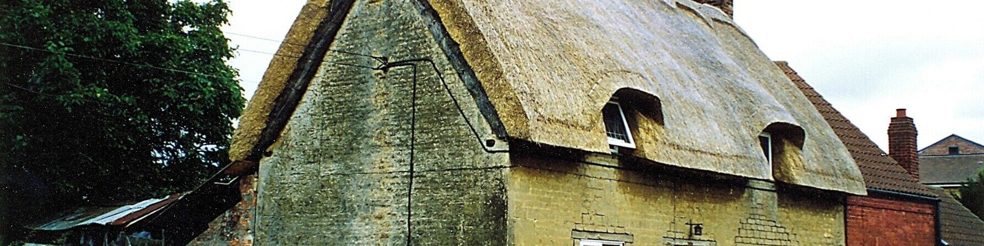 A traditional thatched-roof house with a stone foundation, set in a rural area with a road in the foreground and greenery around.