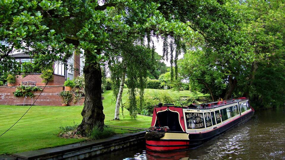 A narrowboat docked by a brick wall with a grassy lawn and trees.
