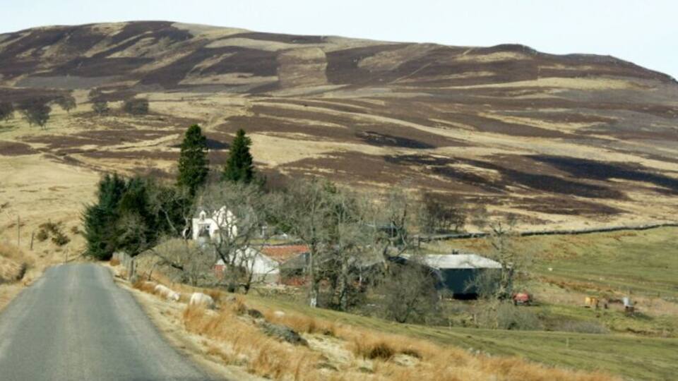 A rural road with a white house and barn in the distance, surrounded by rolling hills and sparse vegetation.