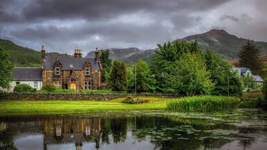 A stone house with a pond in front, surrounded by lush greenery and mountains in the background.