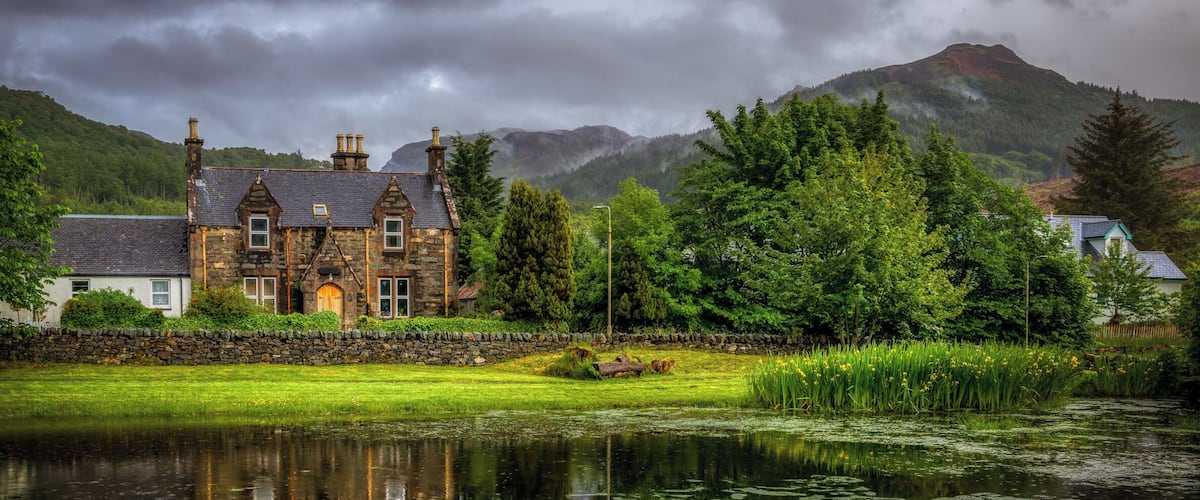 A stone house with a pond in front, surrounded by lush greenery and mountains in the background.