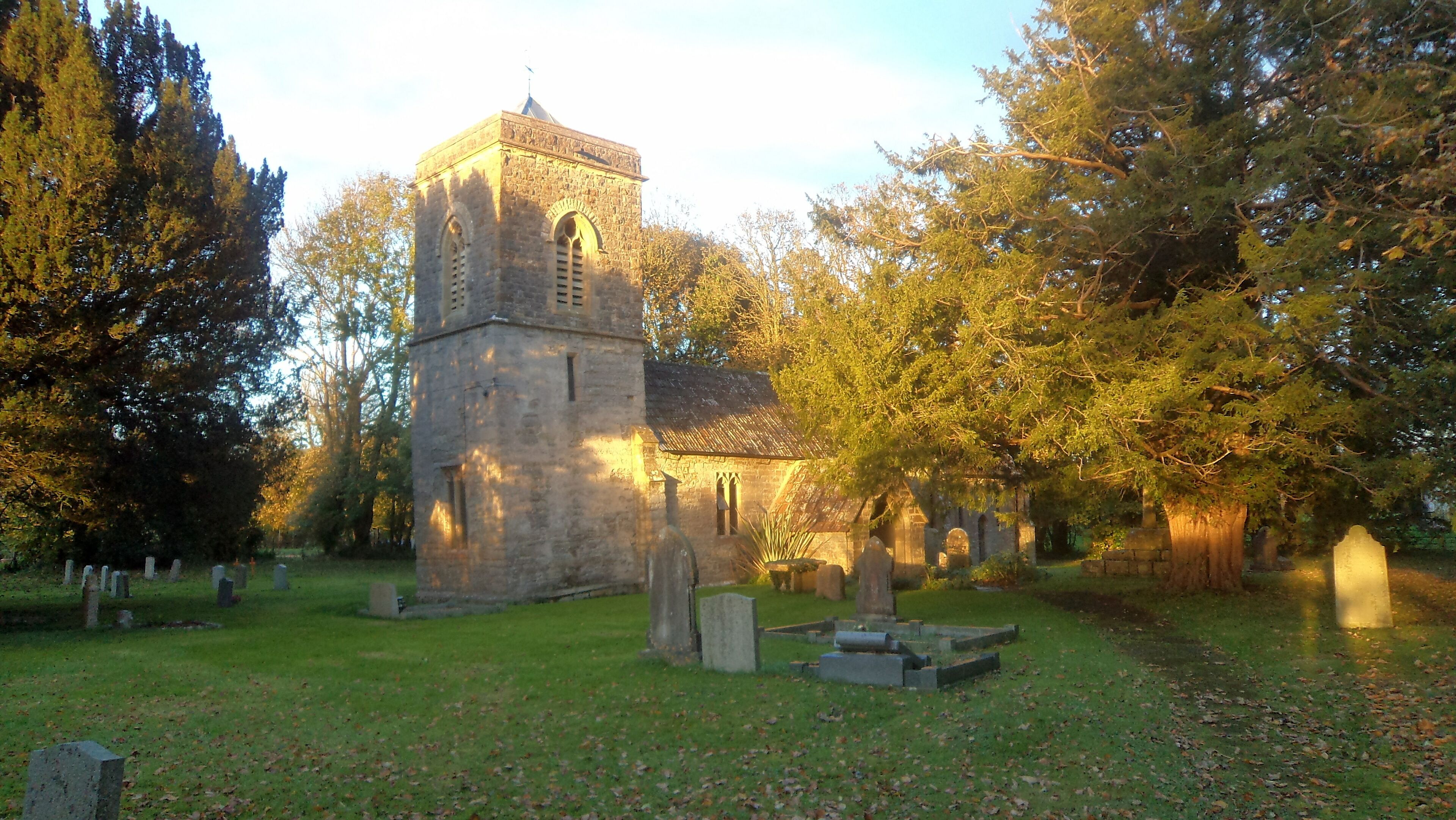 A church with a tall tower and arched windows, surrounded by trees and gravestones in a cemetery.