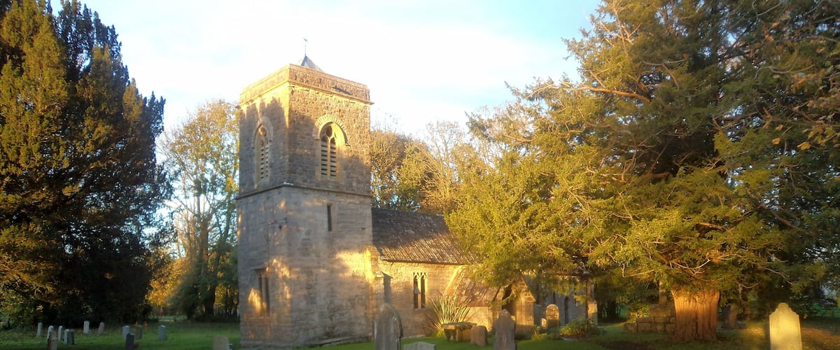 A church with a tall tower and arched windows, surrounded by trees and gravestones in a cemetery.