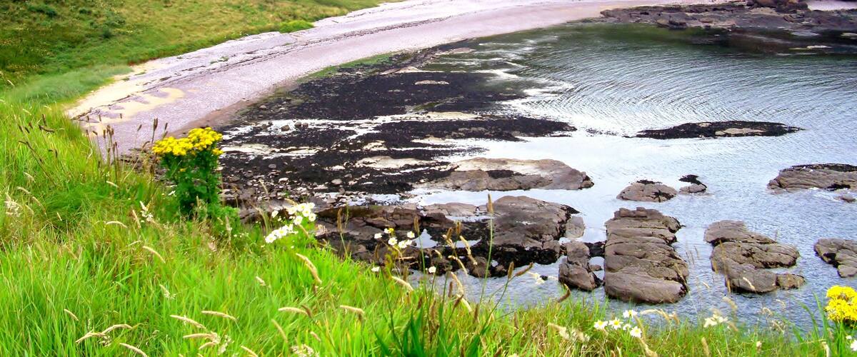 A coastal landscape with a curved sandy beach, rocky outcrops, and lush green hills.