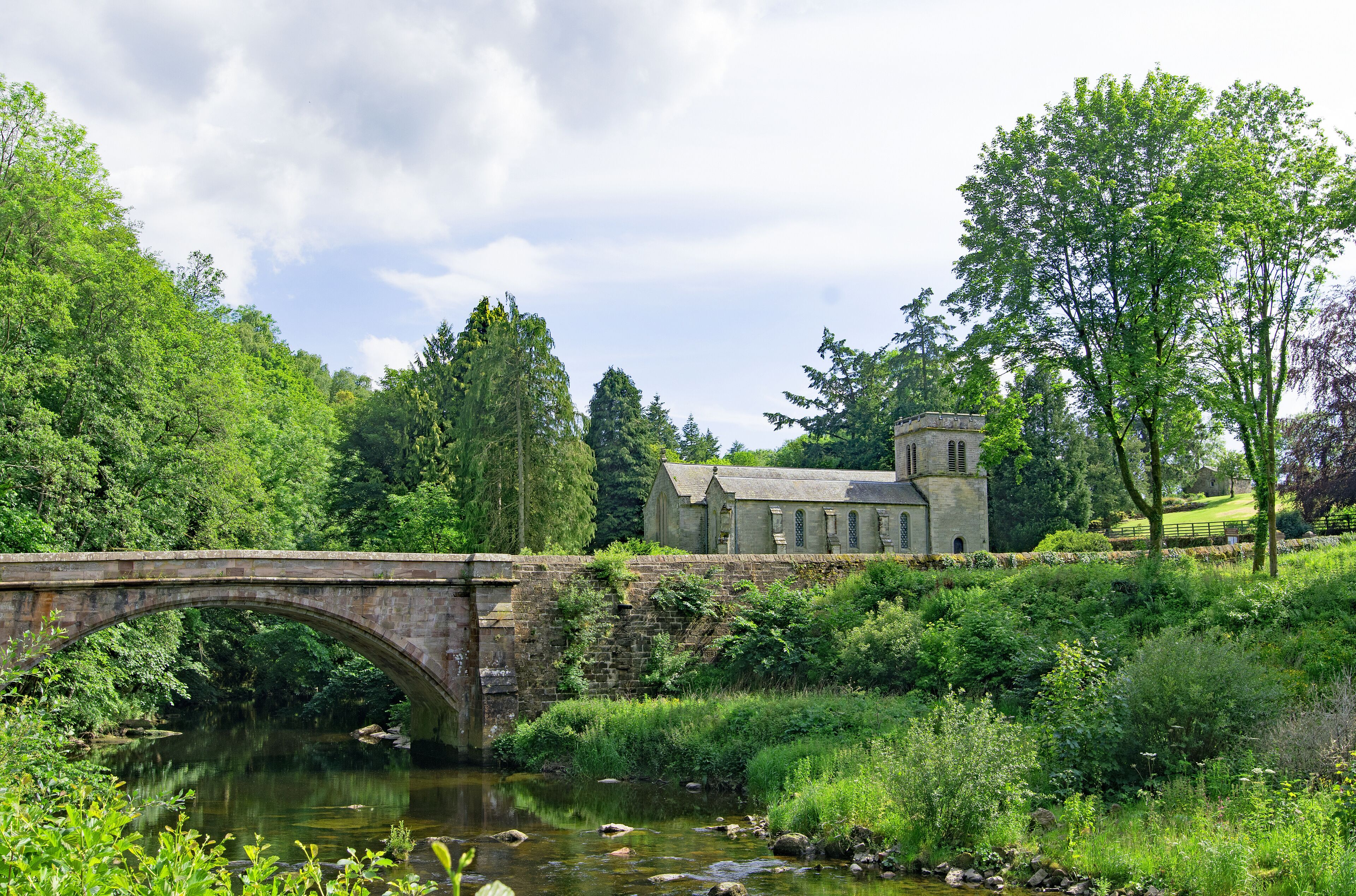 A stone bridge over a river, surrounded by lush greenery and a historic building.
