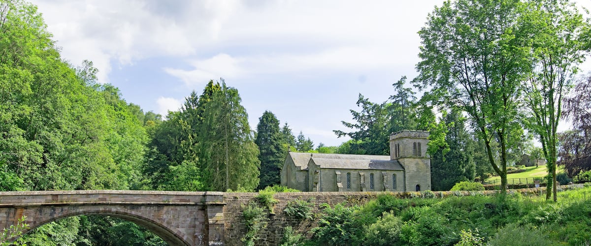 A stone bridge over a river, surrounded by lush greenery and a historic building.