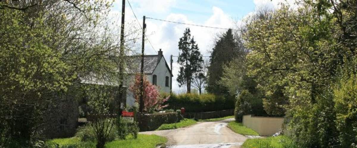 A rural road with puddles, surrounded by greenery and a stone house.