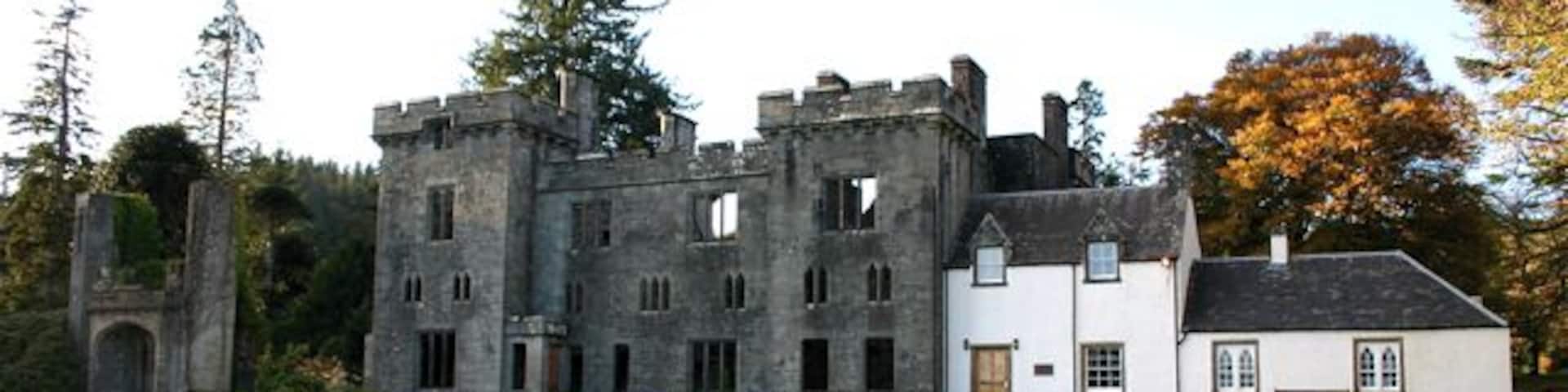 A large, historic stone castle with a white building adjacent to it, set in a grassy field with trees in the background.