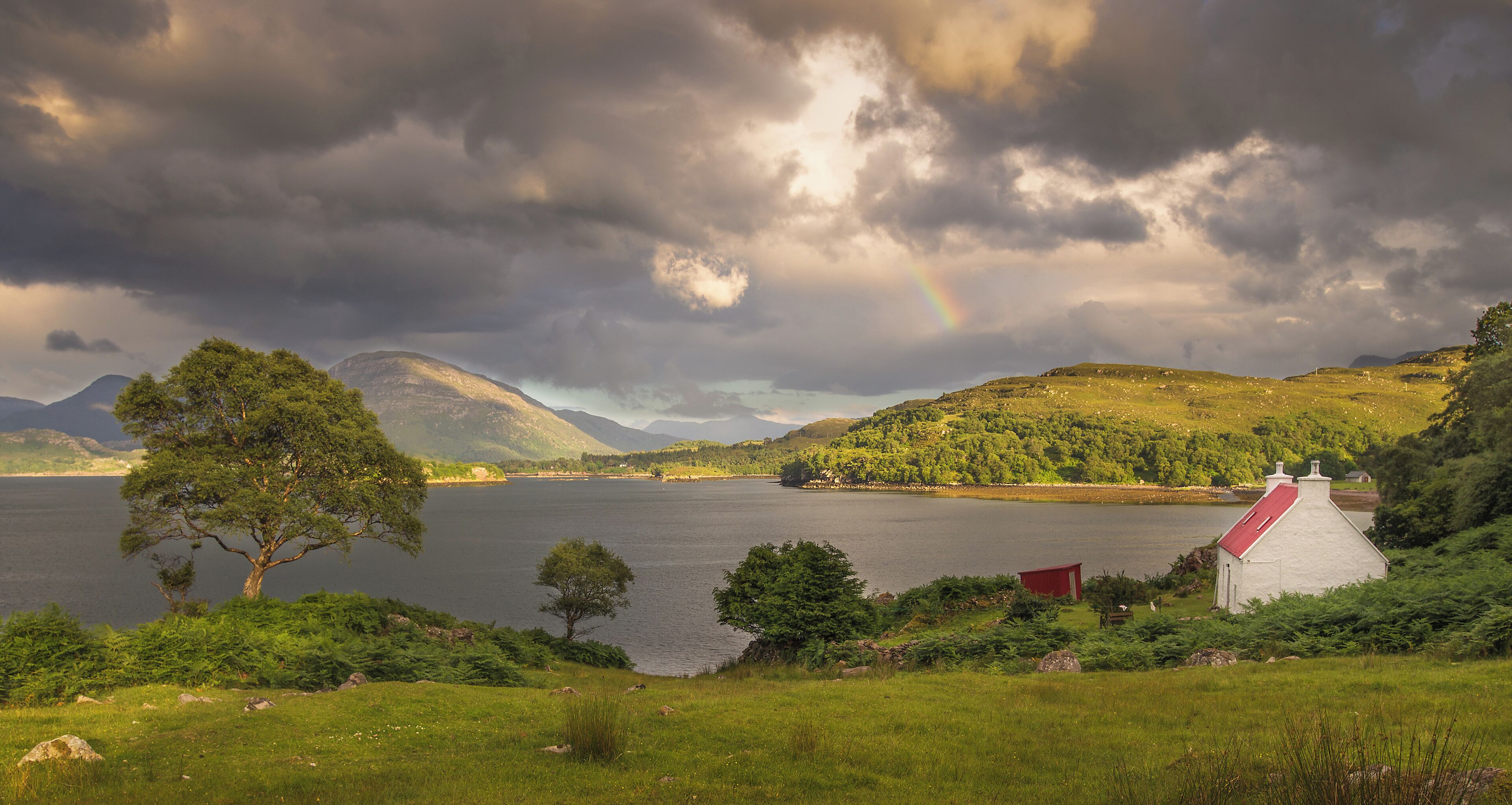 A lake with a rainbow, a red-roofed building, and green hills.