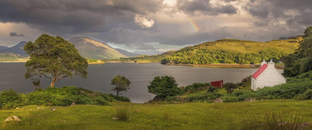 A lake with a rainbow, a red-roofed building, and green hills.