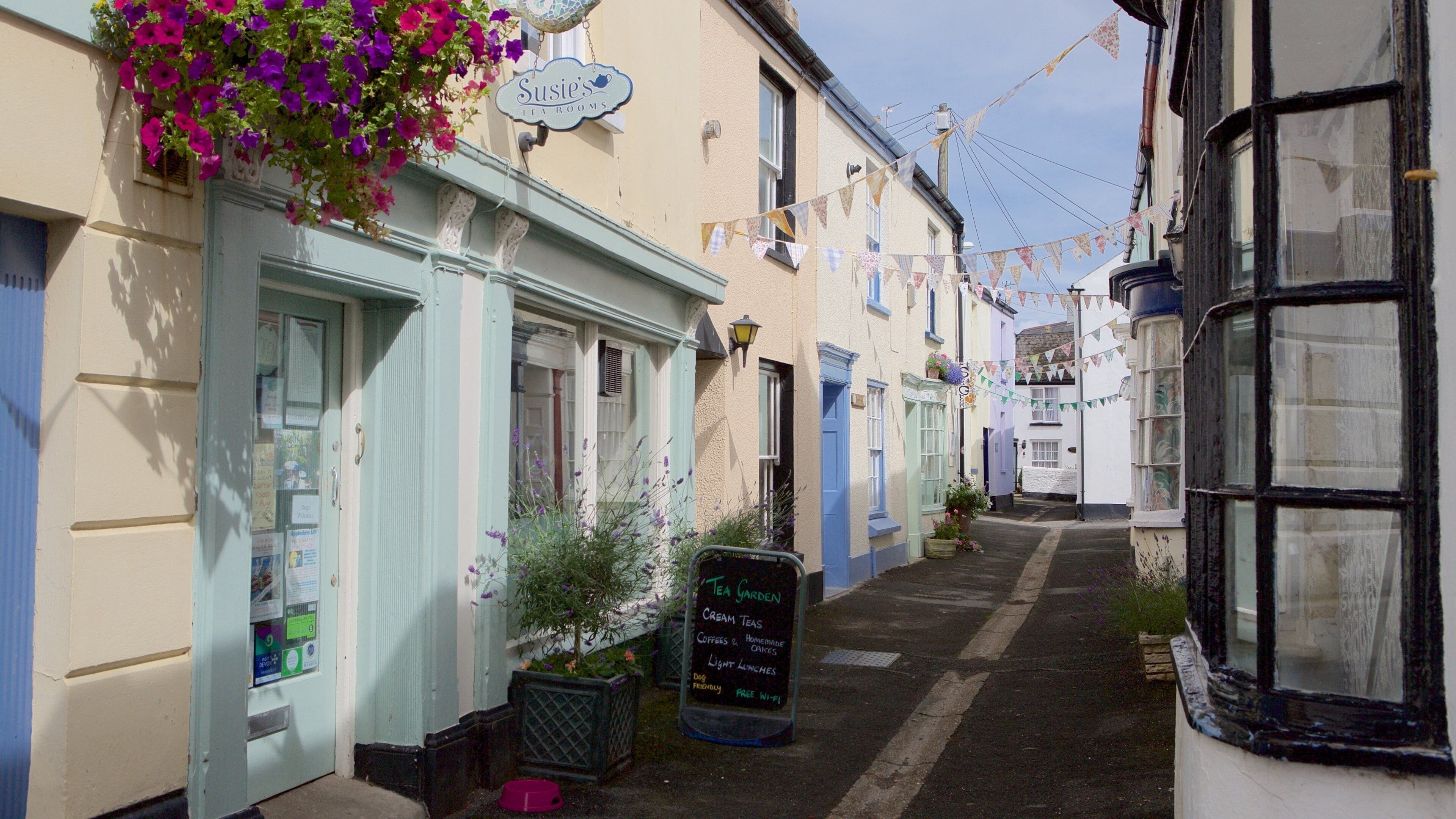 A narrow street with tea shops, a sign for 'Susie's Tea Rooms', and a chalkboard advertising tea garden, cream teas, and light lunches.