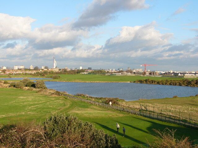 A golf course with a water body, a lighthouse, and industrial buildings in the background.