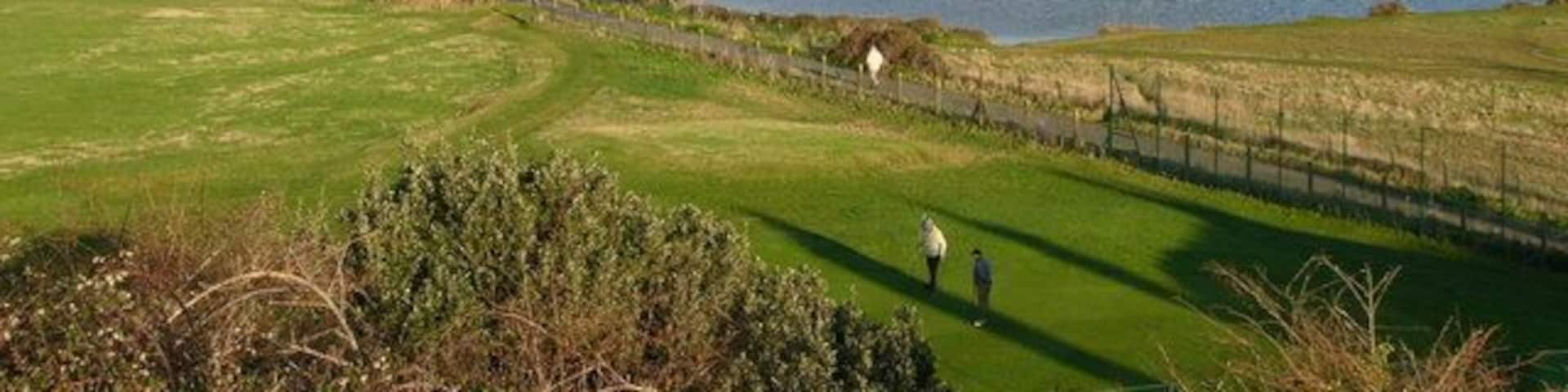 A golf course with a water body, a lighthouse, and industrial buildings in the background.