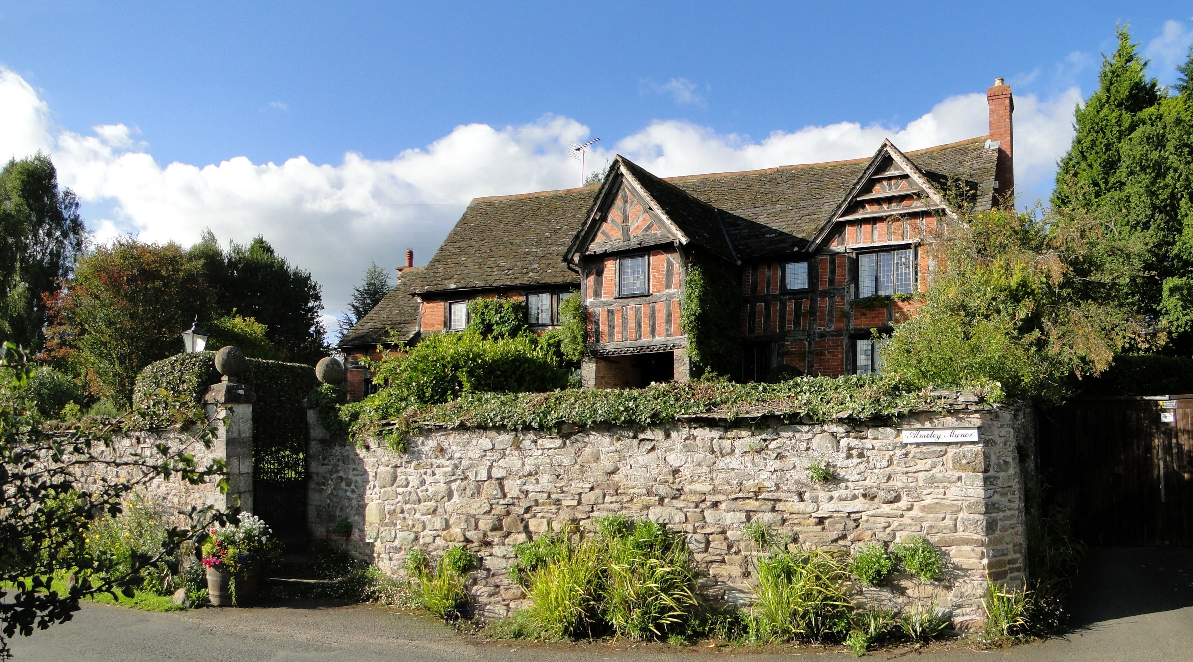 A traditional stone wall with a gate, flanked by lush greenery and a stone lamp post.