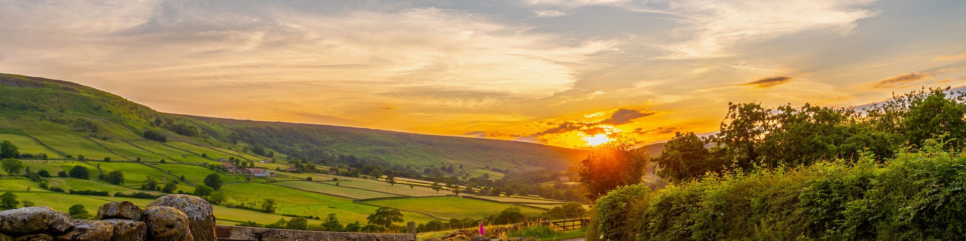 A rural landscape with a stone gate, a dirt road, rolling green hills, and a sunset.