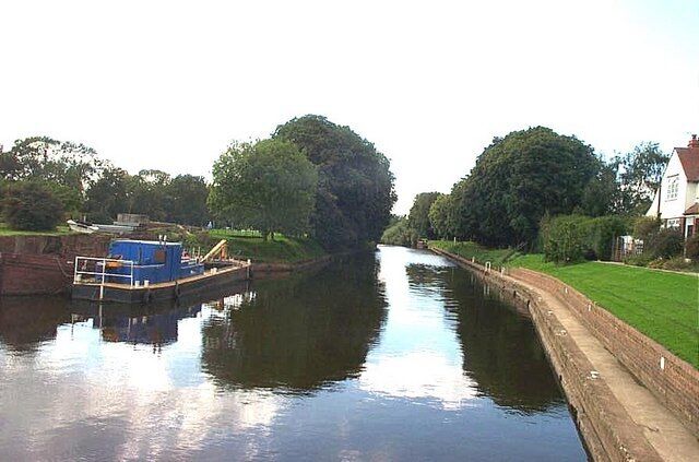 A narrow canal with a blue boat docked on the left, surrounded by greenery and a house on the right.