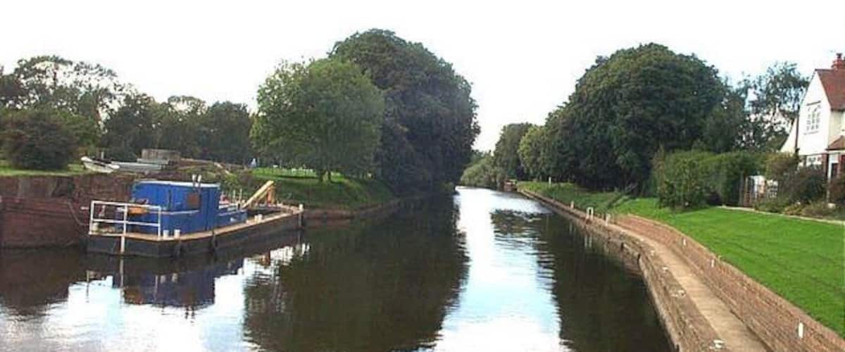 A narrow canal with a blue boat docked on the left, surrounded by greenery and a house on the right.