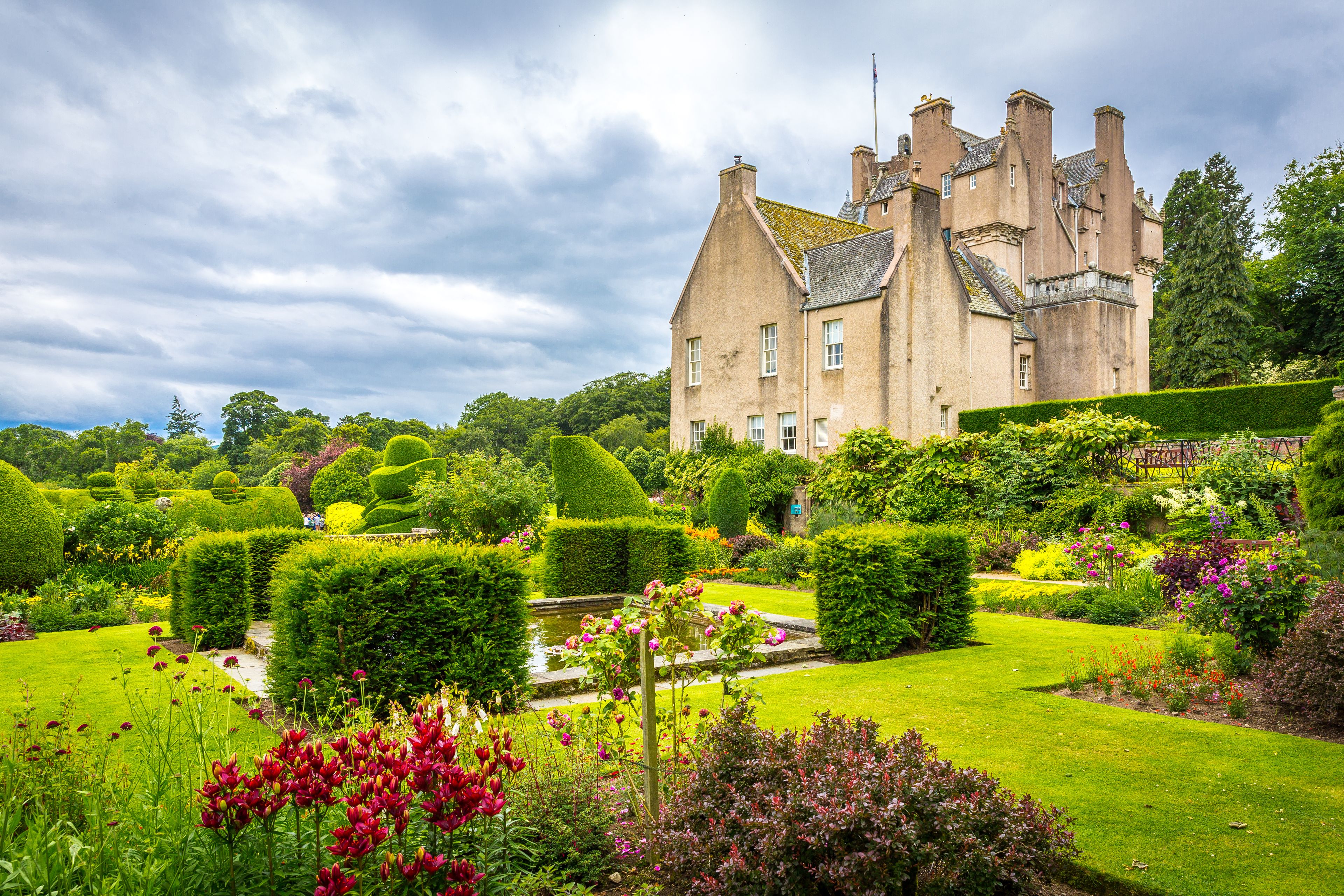 A historic building with a well-manicured garden, including hedges, flowers, and a pond.