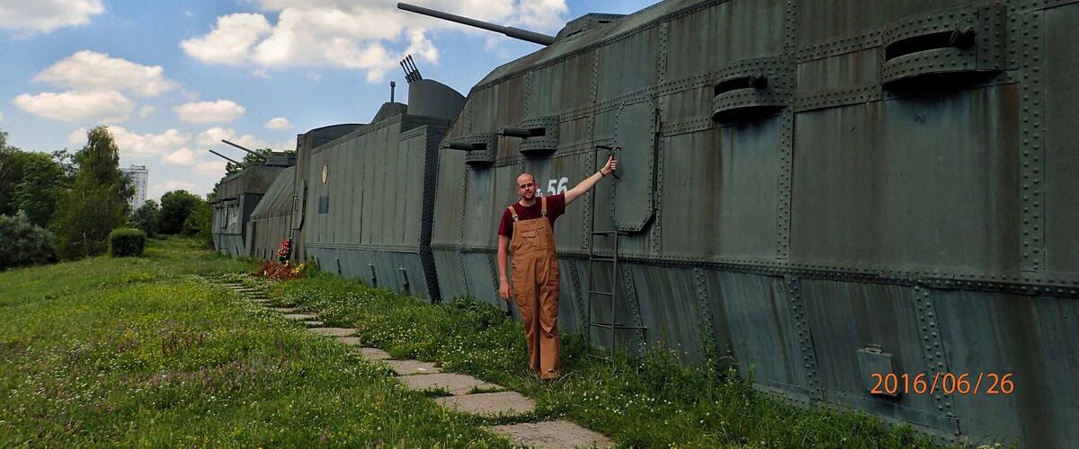 A person standing next to a large, green military tank with a cannon, with a clear sky and greenery in the background.